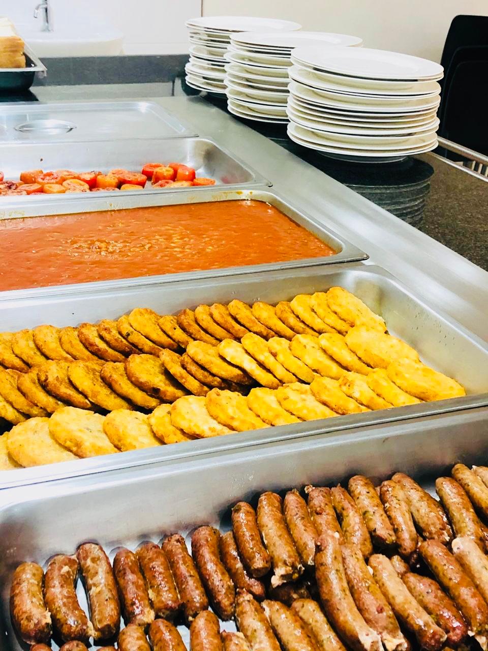 A hot breakfast line of boerewors, fritters, baked beans and grilled tomato with stacked service plates.