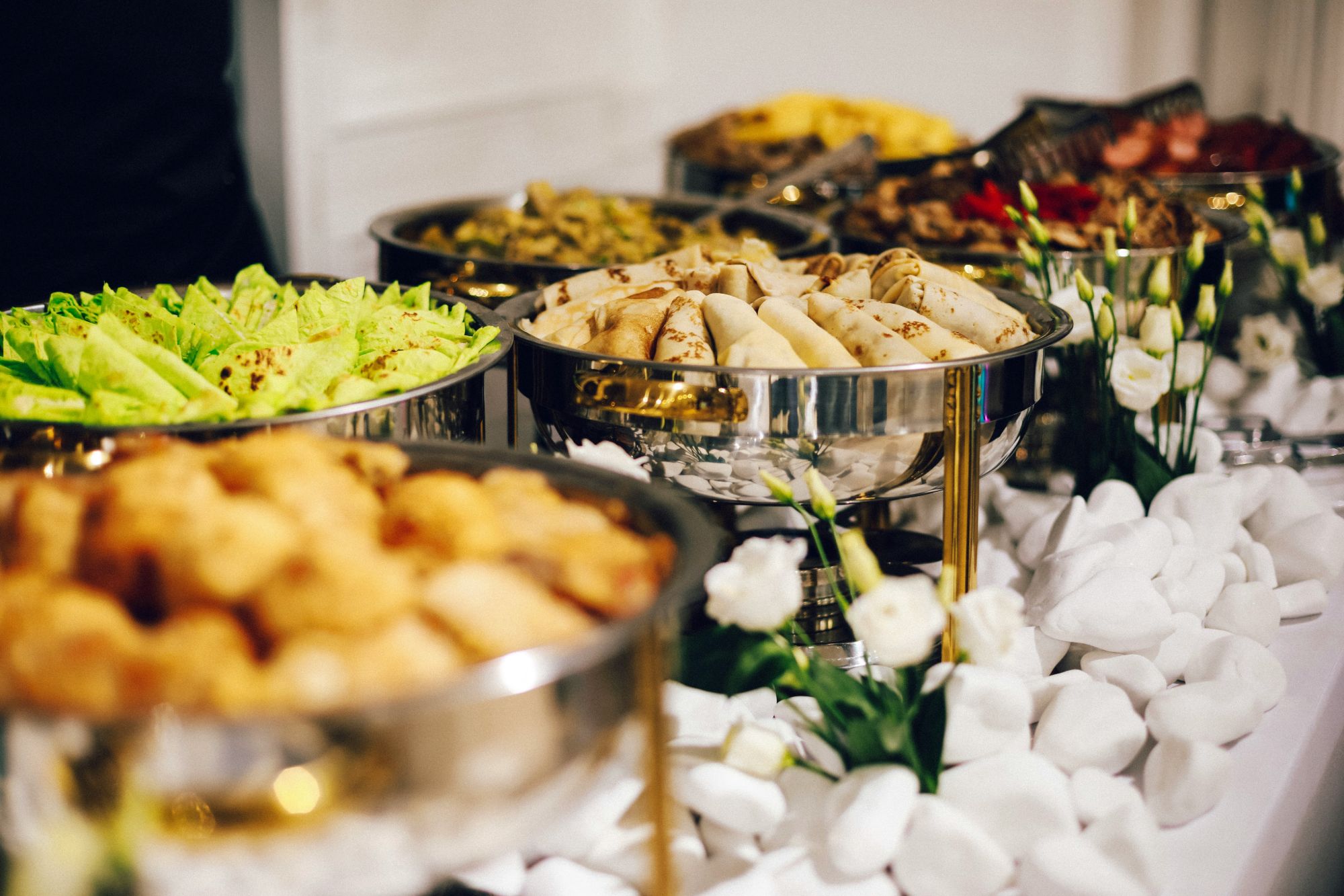 An event buffet with silver serving bowls of wraps, salad, pastries and hot dishes styled with white florals.