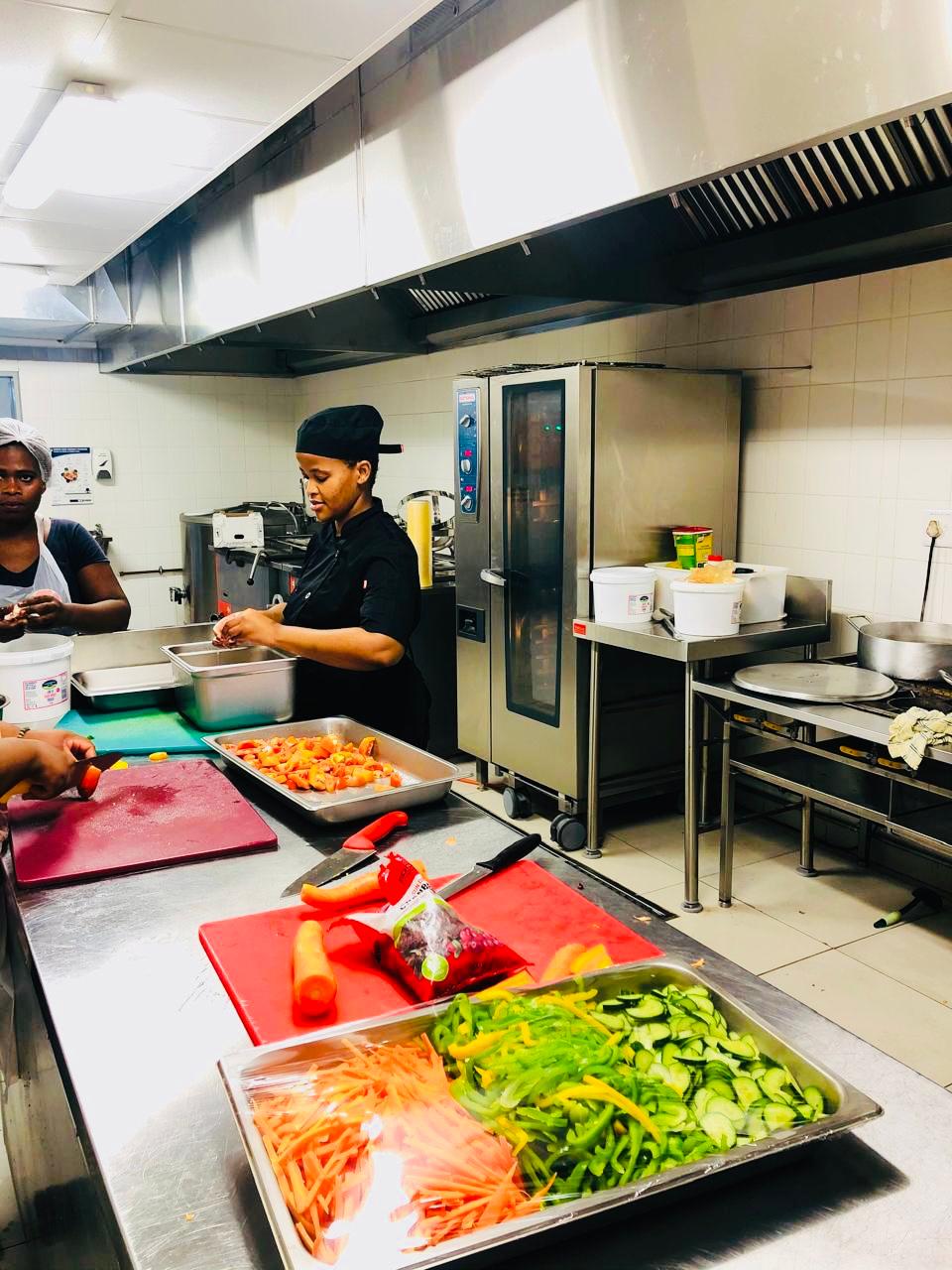 Abaholi kitchen team preparing trays of julienned vegetables in a stainless steel production kitchen.
